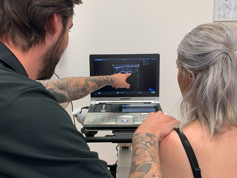 Een man bij Fysio Medisch Centrum Rijssen laat een vrouw met grijs haar een echo op een laptop zien, terwijl hij naar het scherm wijst.